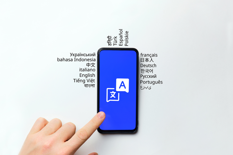 Image of a left hand pointing at an iPhone screen with languages displayed all around the iPhone. The image showcases a hand holding an iPhone, with the screen showing content related to languages. Surrounding the iPhone are icons, symbols, or text representing different languages, indicating a diverse linguistic environment. The hand pointing at the screen suggests interaction and engagement with language-related content on the iPhone. The image symbolizes the use of technology for accessing multilingual resources, language learning, or communication in various languages.